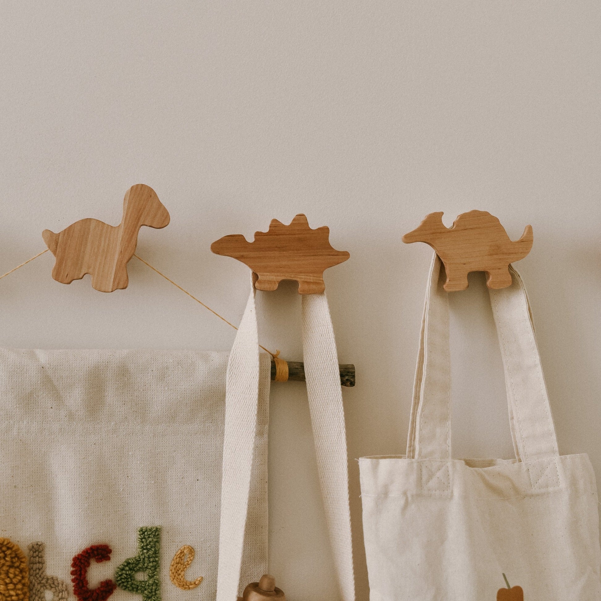 Three wooden dinosaur-shaped hooks on a wall with fabric bags hanging from them.