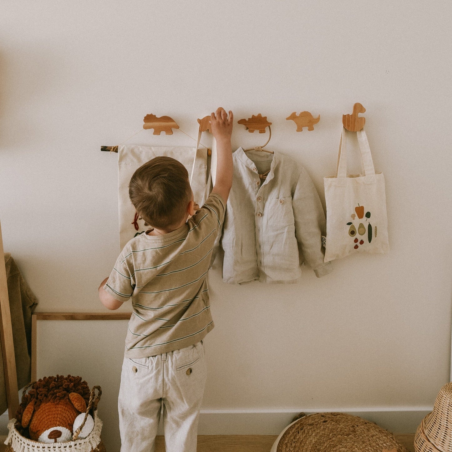Child hanging a shirt on a hook in a room with a basket of toys and a bag.