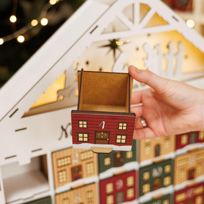 Hand holding a small red house-shaped box in front of a decorative Advent calendar with lit lights.