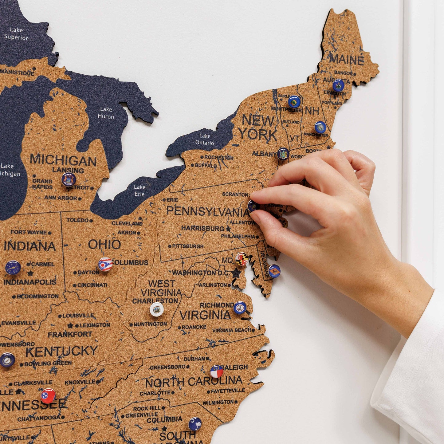 Hand placing a pin on a corkboard map of the United States with pins marking various locations.