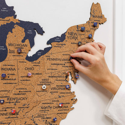 Hand placing a pin on a corkboard map of the United States with pins marking various locations.