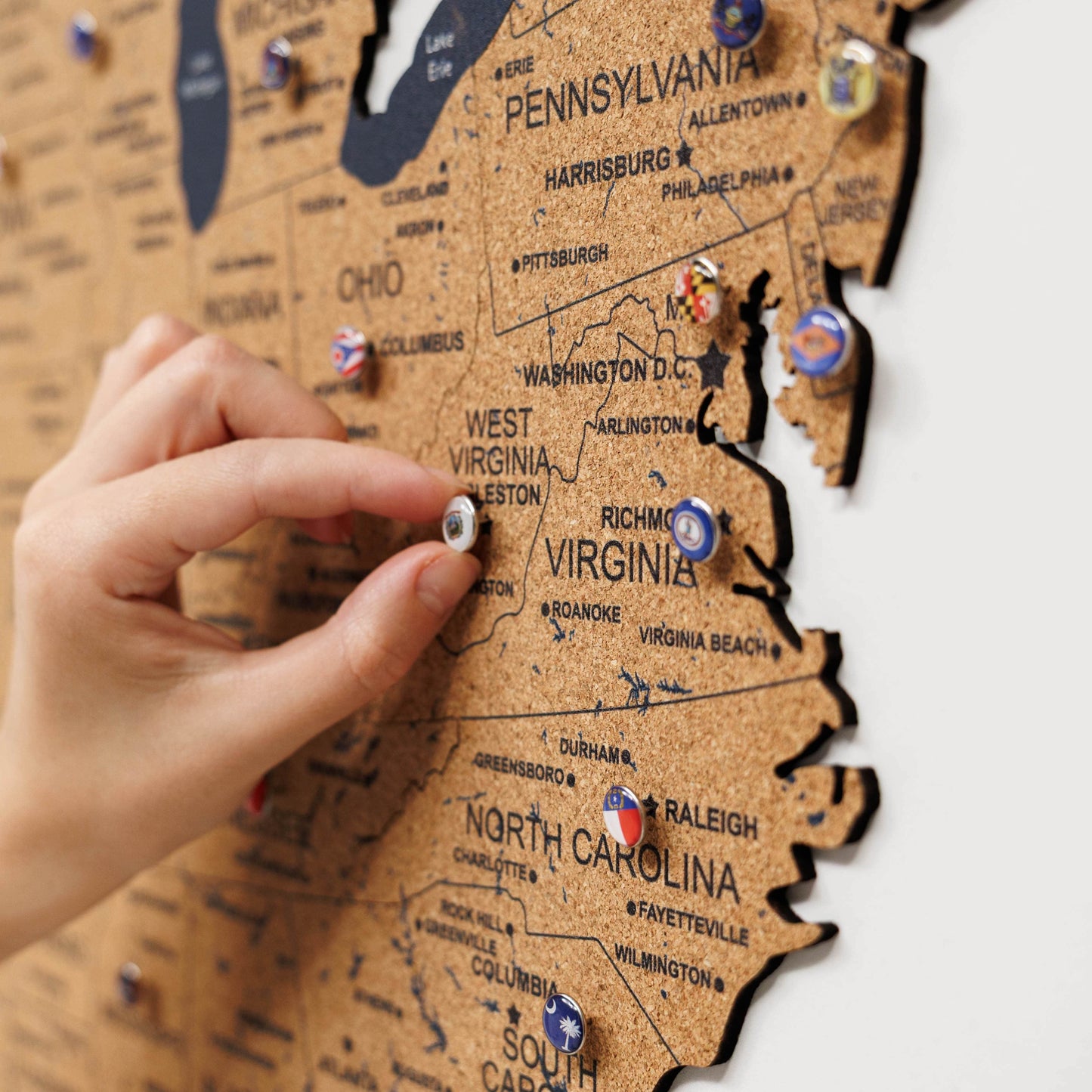 Hand placing a pin on a wooden map of the United States with pins marking various locations.