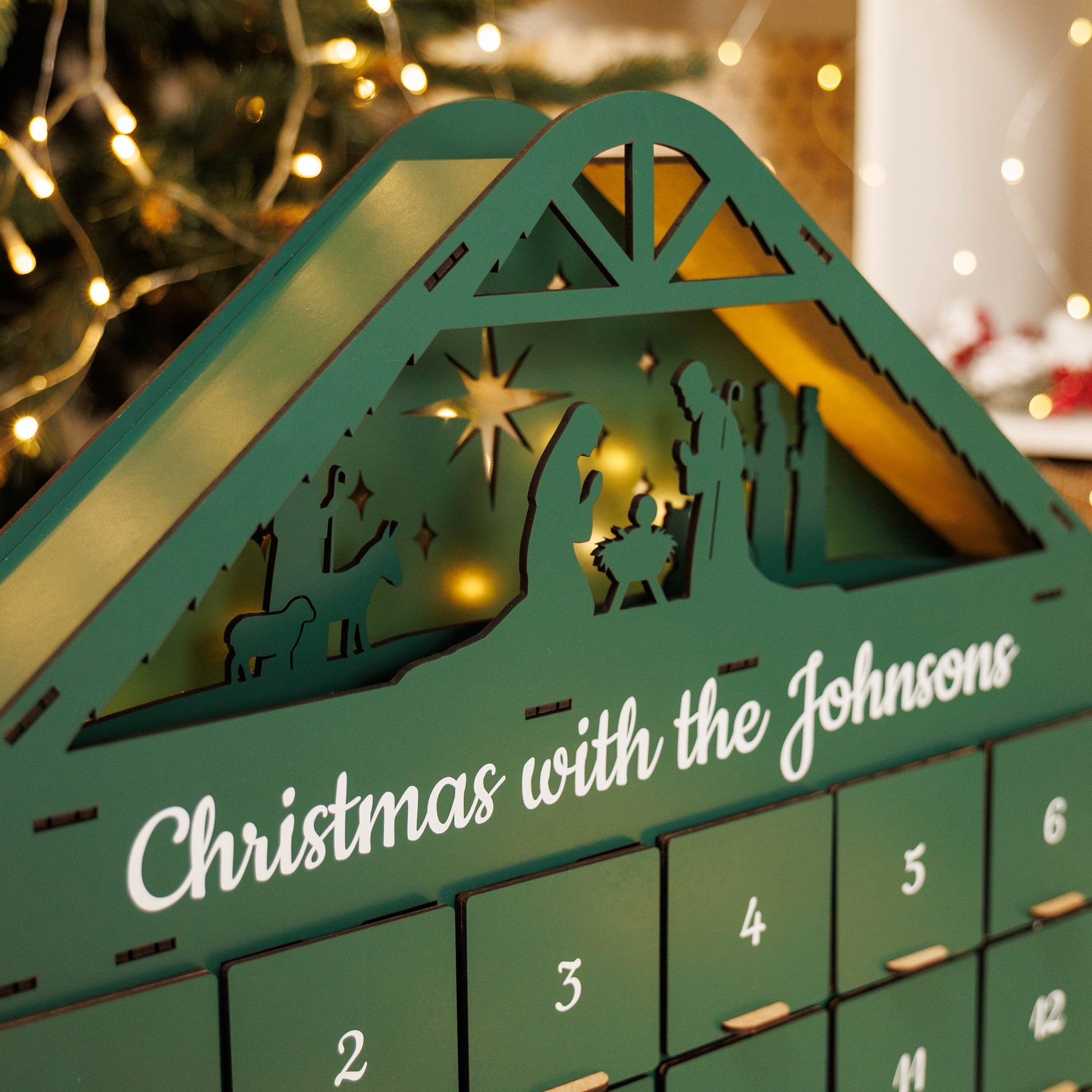 Advent calendar with nativity scene design and 'Christmas with the Johnsons' text, blurred Christmas lights in the background.