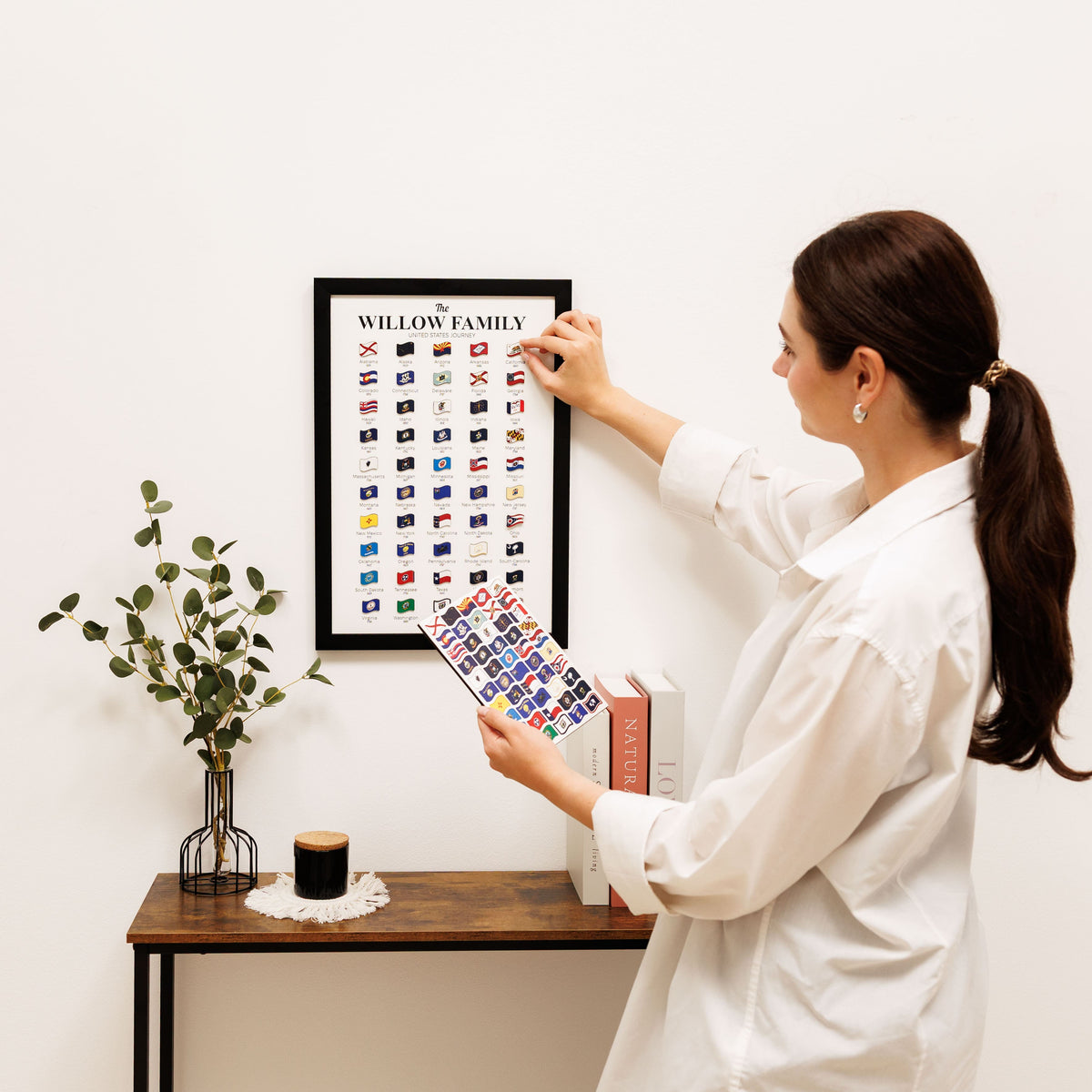 Woman hanging a framed print on a wall next to a small table with decor items.