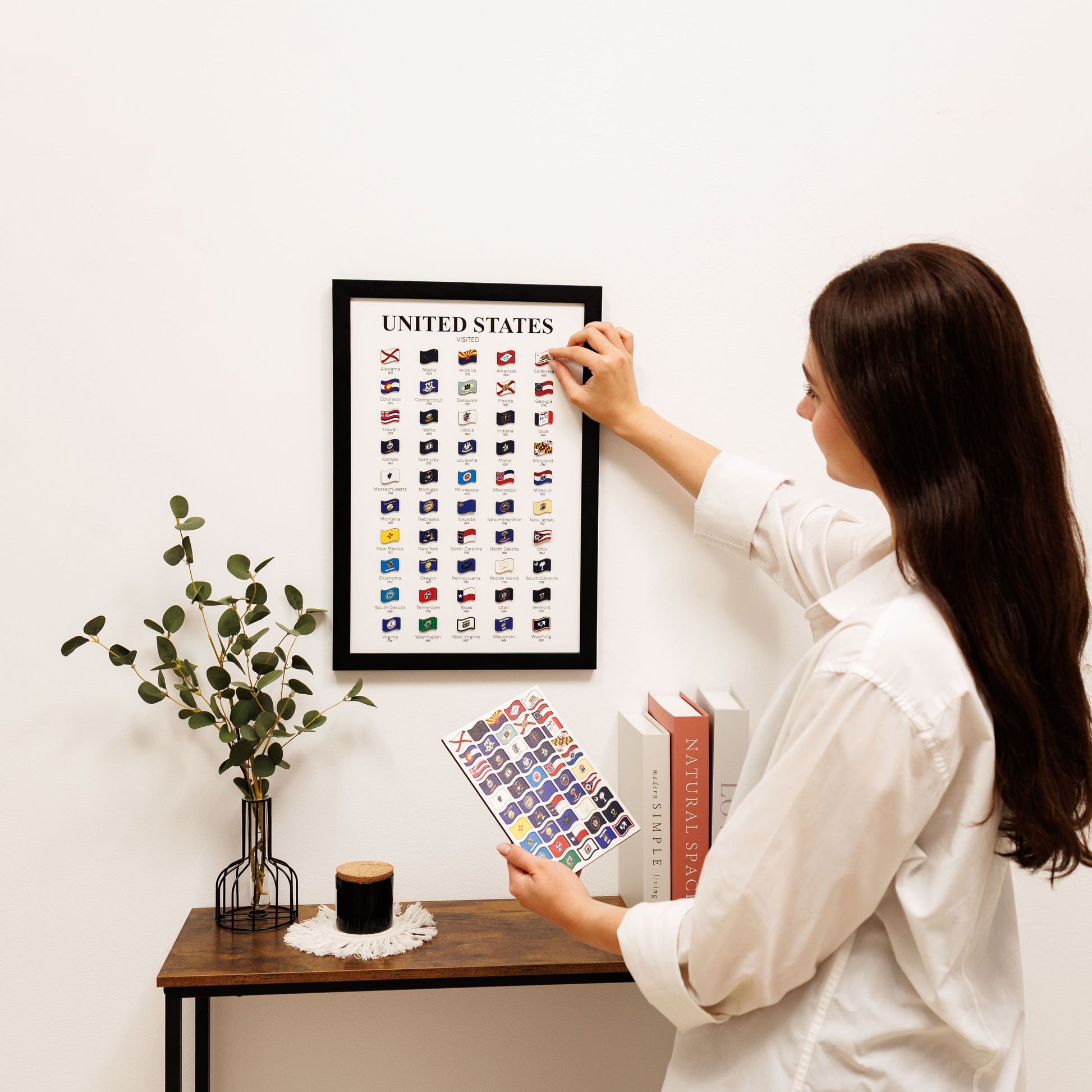 Woman hanging a framed print of flags on a wall next to a table with decor items.