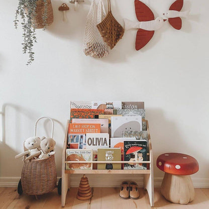 A wooden Montessori bookshelf designed for children, featuring a low shelf packed with books, placed against a white wall in a home setting. The bookshelf is made of natural wood and has a front-facing design to display book covers.