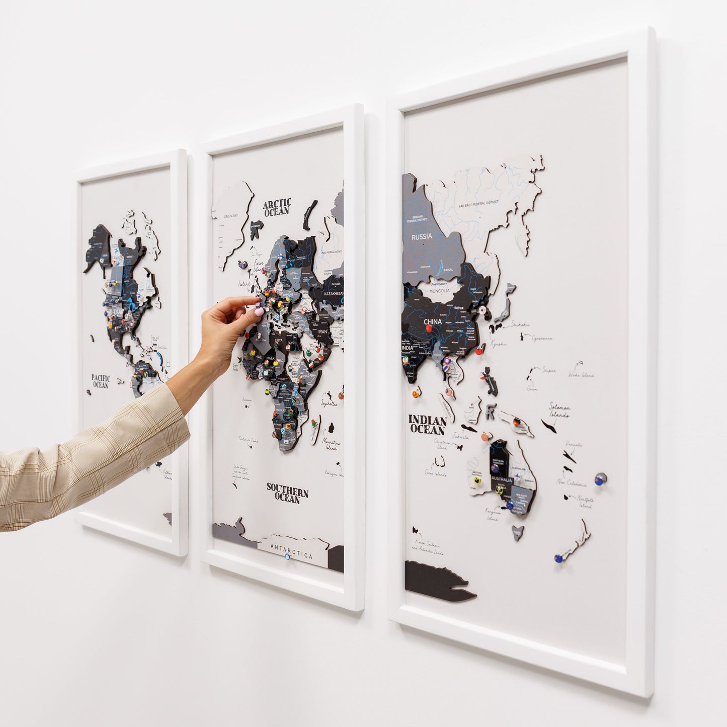 Three framed world maps with pins on a white wall, one map is being interacted with by a hand.