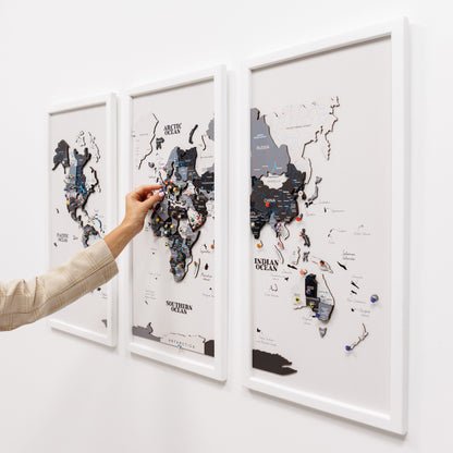 Three framed world maps with pins on a white wall, one map is being interacted with by a hand.
