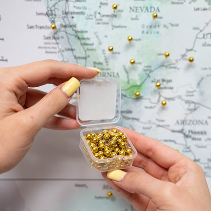 Hand holding a small container of gold beads with a map of the United States in the background.