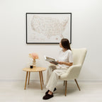 Woman sitting in a chair reading a book in a room with a framed world map on the wall.
