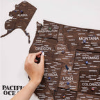 Wooden map of the United States with pins marking locations, held by a hand.