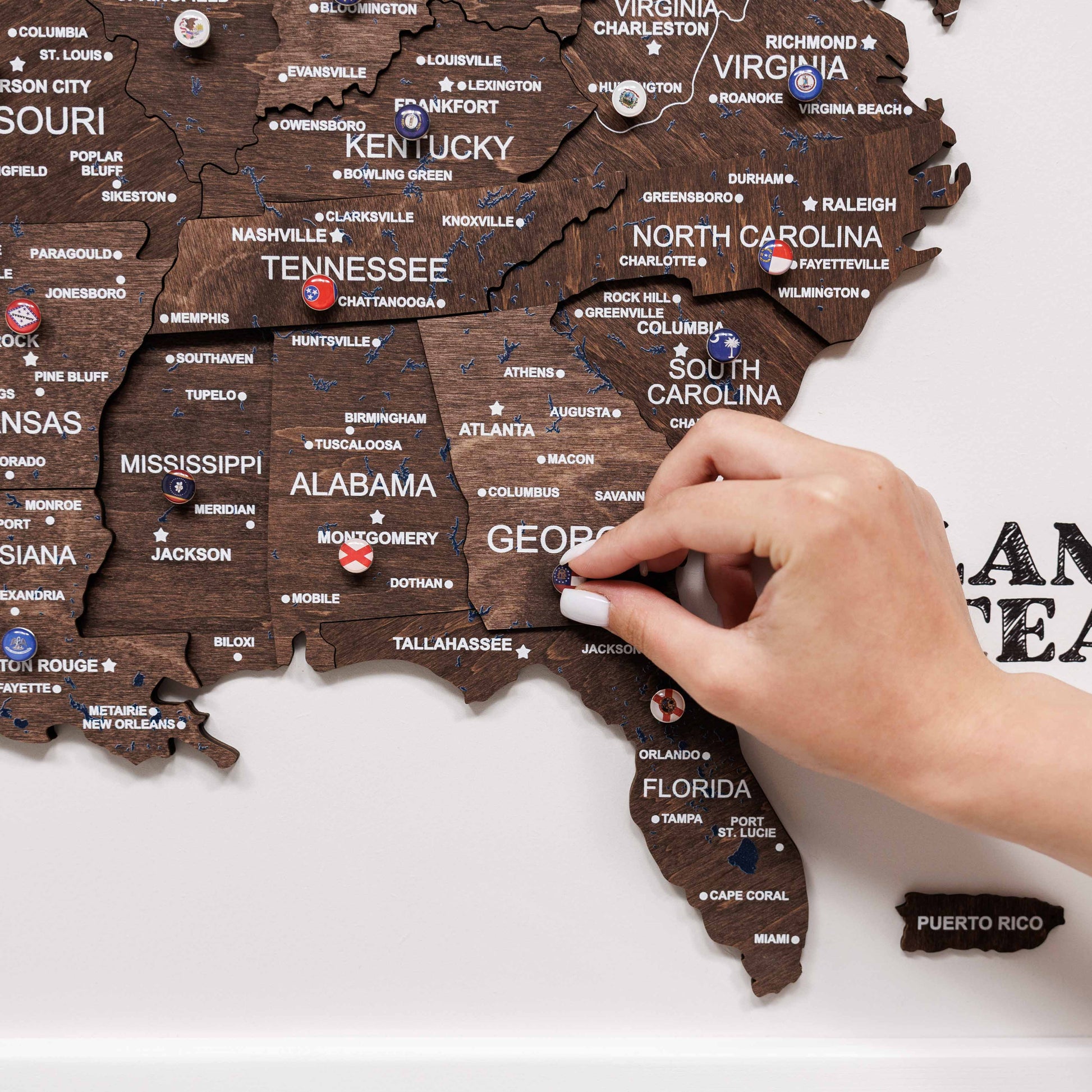 Wooden map of the United States with a hand placing a red pin on Alabama.