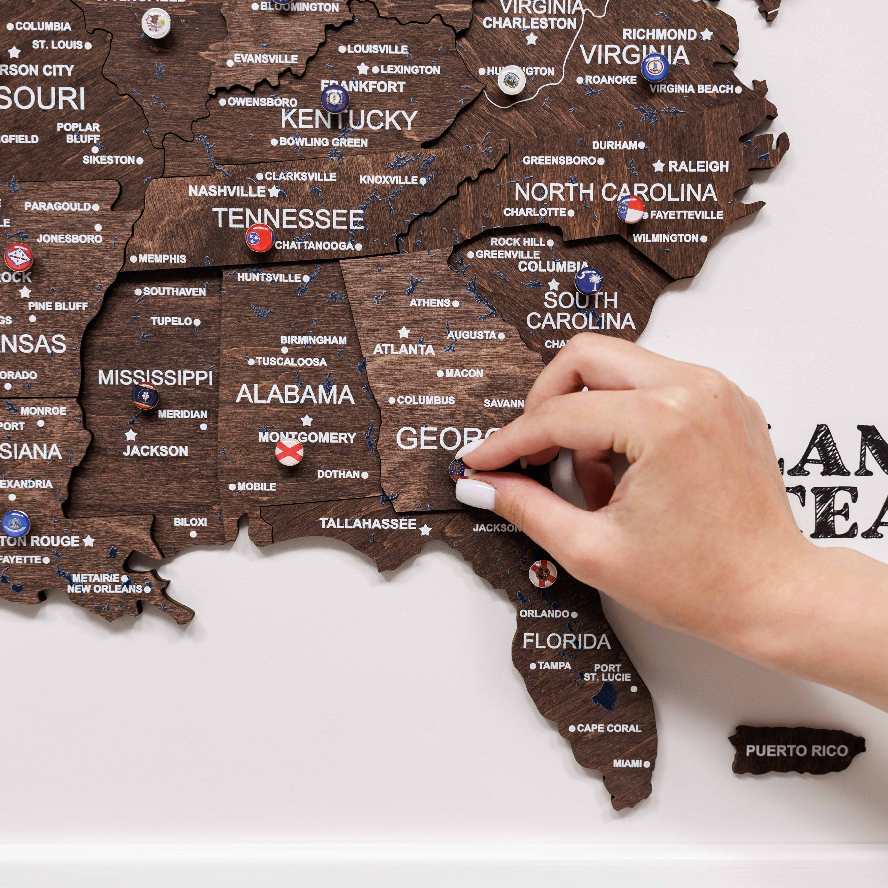 Wooden map of the United States with a hand placing a red pin on Alabama.