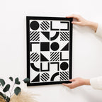 Person holding a framed black and white geometric art print on a light surface.