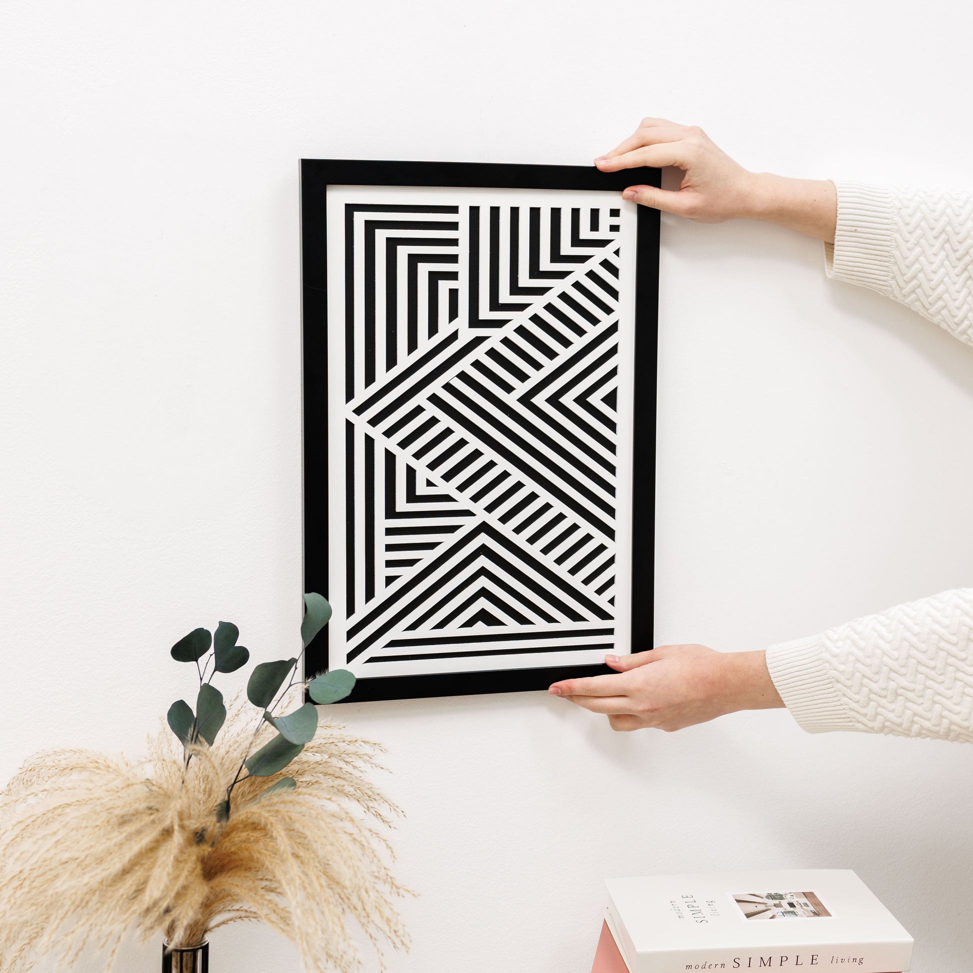 Person holding a black frame with a geometric pattern on a white surface