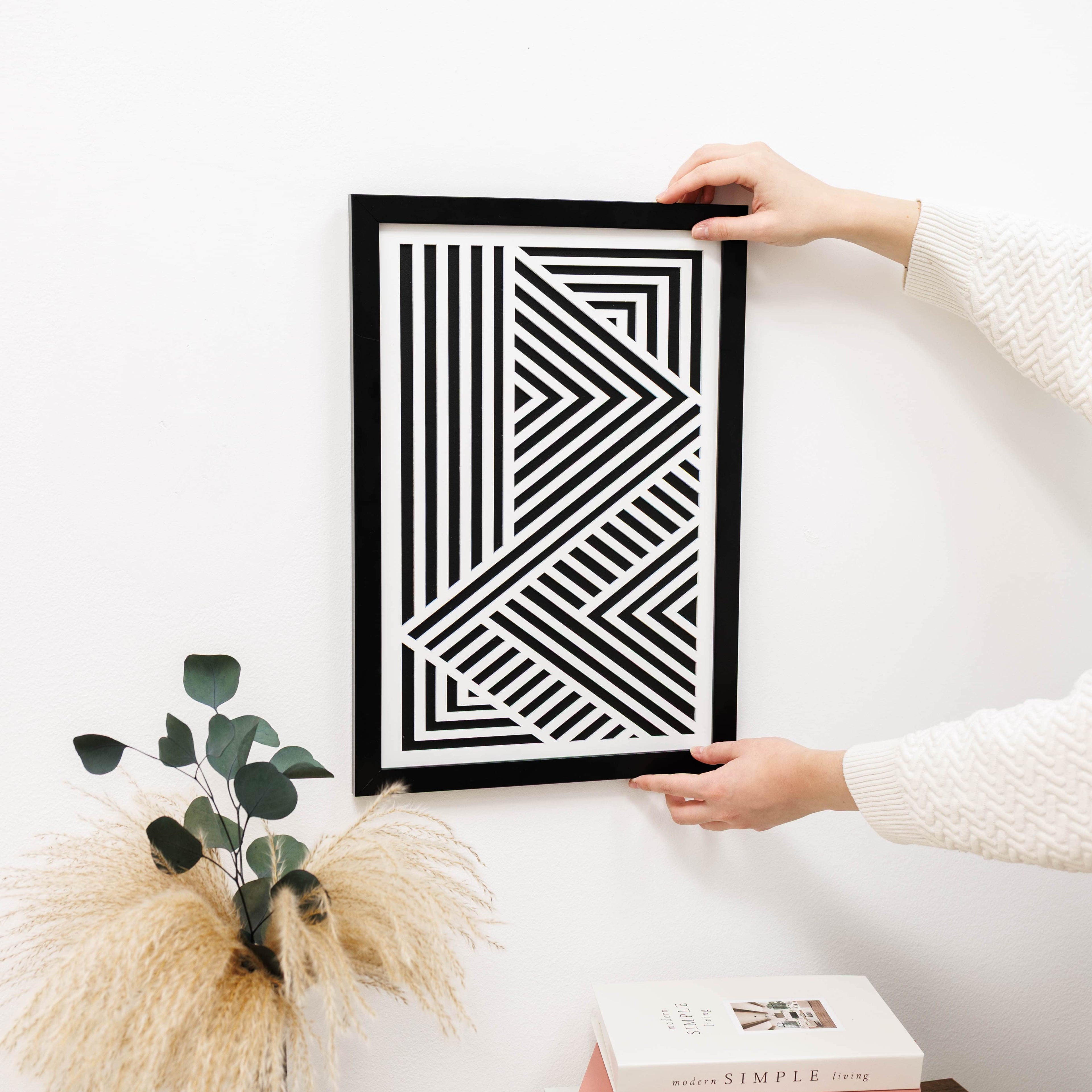 Person holding a black frame with a geometric design on a white wall.