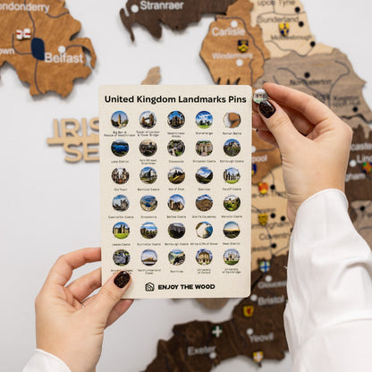 Card with United Kingdom landmarks pins held by a person in front of a wooden map.